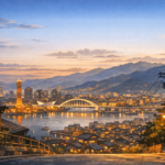 A quiet, wide view of Kobe from a hillside at dusk, with soft city lights, the harbor in the distance, and mountains fading into the evening.