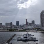 A calm waterfront view of Kobe Harborland and Meriken Park under cloudy skies, with sunlight breaking through the clouds and reflecting on the sea.