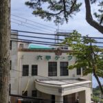 The historic Art Deco building of Rokko Cable Sanjo Station, framed by tall pine trees under a clear sky.