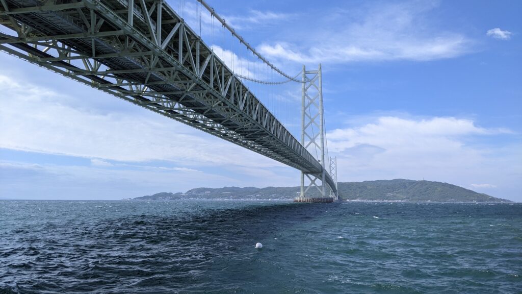 A powerful low-angle view of the Akashi Kaikyo Bridge's white steel structure stretching across the deep blue sea toward Awaji Island under a bright sky.