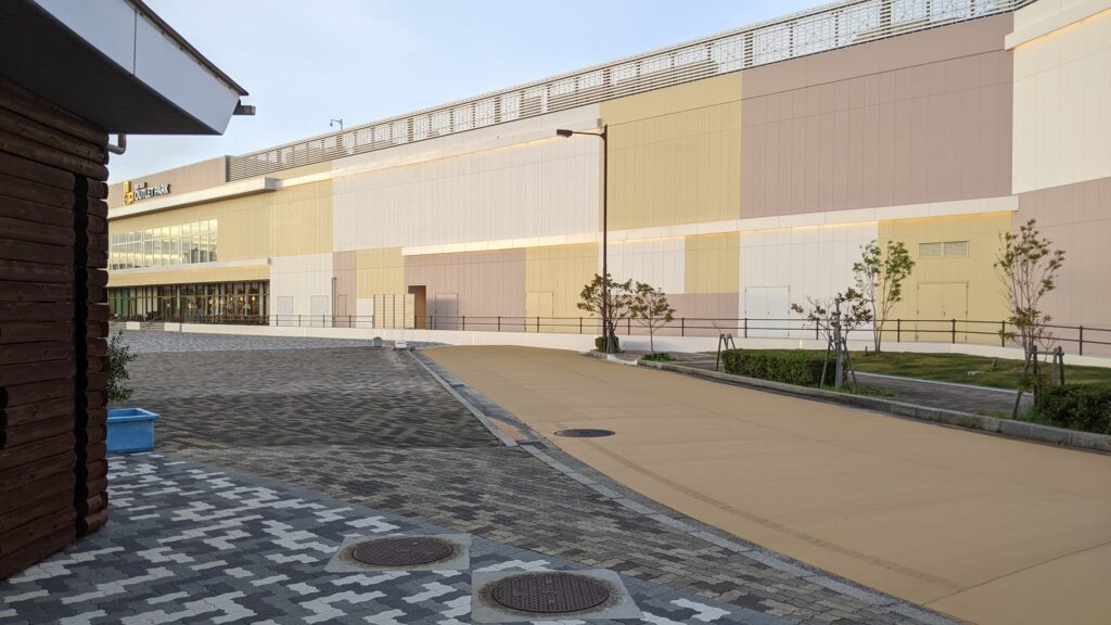A modern and colorful facade of Mitsui Outlet Park Marine Pia Kobe with wide paved walkways and young trees under a clear sky.
