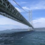 A powerful low-angle view of the Akashi Kaikyo Bridge's white steel structure stretching across the deep blue sea toward Awaji Island under a bright sky.