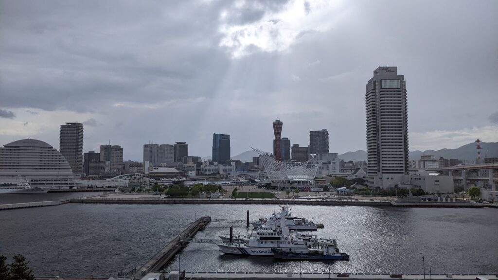 A serene waterfront view of Kobe's Harborland and Meriken Park. Sunlight breaks through the clouds over the sea, illustrating the "quiet rhythm" of the city that inspires the kodiad project.