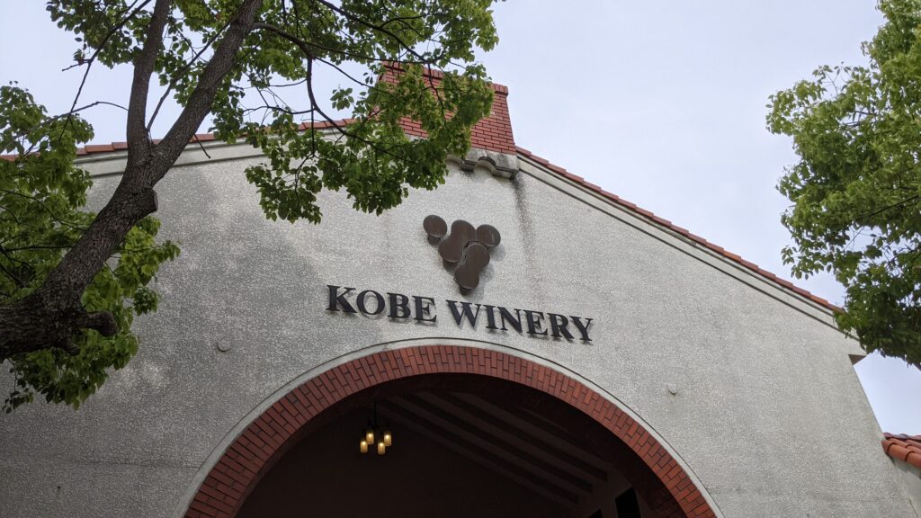The brick arch entrance of Kobe Winery (Aguri Park) under a bright sky, surrounded by green trees. The sign "KOBE WINERY" is visible on the wall.