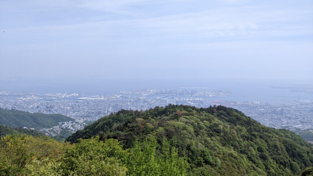 A panoramic view of the Kobe metaverse inspiration from Mt. Rokko Observatory, showing the "vertical rhythm" between the lush green mountains and the city skyline under a clear sky.